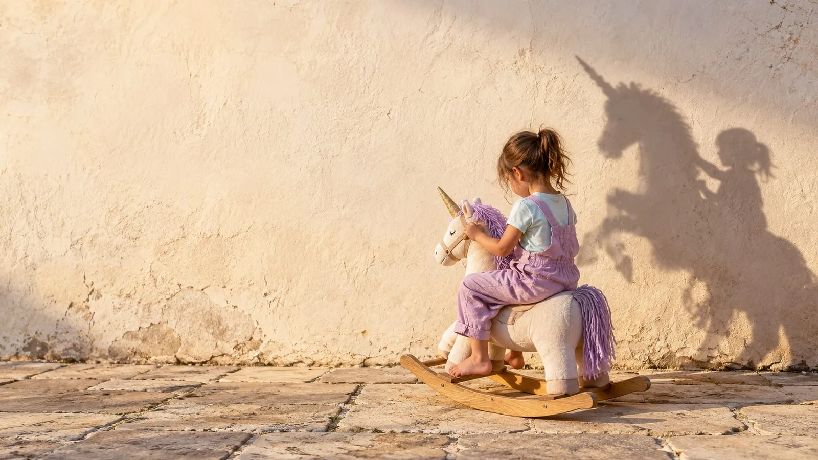Una niña de espaldas monta un unicornio balancín de peluche lila junto a una pared soleada; su sombra en la pared parece un unicornio de verdad